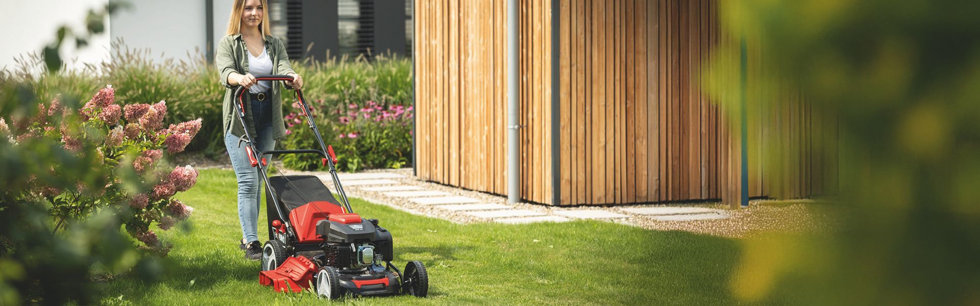 Eine Person mäht mit einem Rasenmäher auf einem grünen Rasen. Im Hintergrund sind Blumen und ein Holzzaun zu sehen.